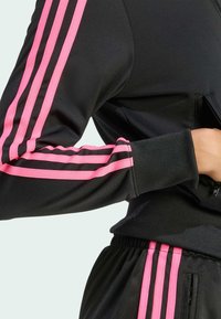 Persona vestita con abbigliamento sportivo nero con strisce rosa sulle maniche e sui pantaloni, mano infilata nella tasca laterale con zip.