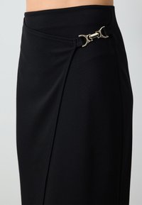Zwarte rok van rekbaar materiaal met een aansluitende taille, voorzien van een diagonale wikkelontwerp en metalen hardware accent bij de taille.