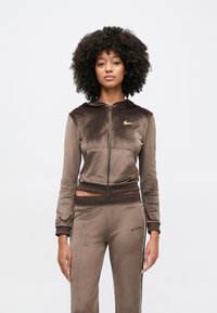 Sweat à capuche zippé en velours marron avec une capuche et des poches avant, assorti à un pantalon de survêtement coordonné. Il présente des accents de logo dorés et des poignets contrastants.