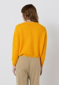 Geltonas trumpintas megztinis pagamintas iš minkštos, tekstūruotos medžiagos, su riboto apdaila apačioje ir rankovėse. Derinamas su smėlio spalvos, laisvo silueto kelnėmis.