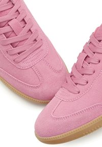 Pink suede sneakers with a smooth texture, featuring matching laces, a rounded toe, and a gum rubber outsole. Simple design and stitching details.