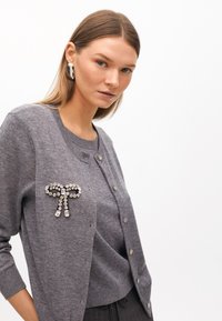 Szara dzianinowa kardigan z dekoracyjną akcentem w postaci kokardy z dżetów. Posiada zapięcia na guziki i okrągły dekolt, zestawiony z pasującą szarością bluzką.