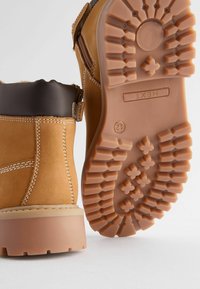 Brun läderstövel med beige accenter, som har en texturerad gummisula och sömnadsdetaljer. Baksidan har ett justerbart band.