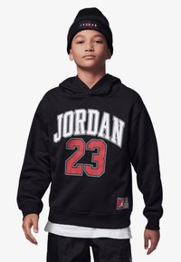 Hoodie preto com o texto branco "JORDAN" e o "23" vermelho na frente. Apresenta um bolso canguru e uma etiqueta com um logótipo na barra. Modelo usando um gorro preto.