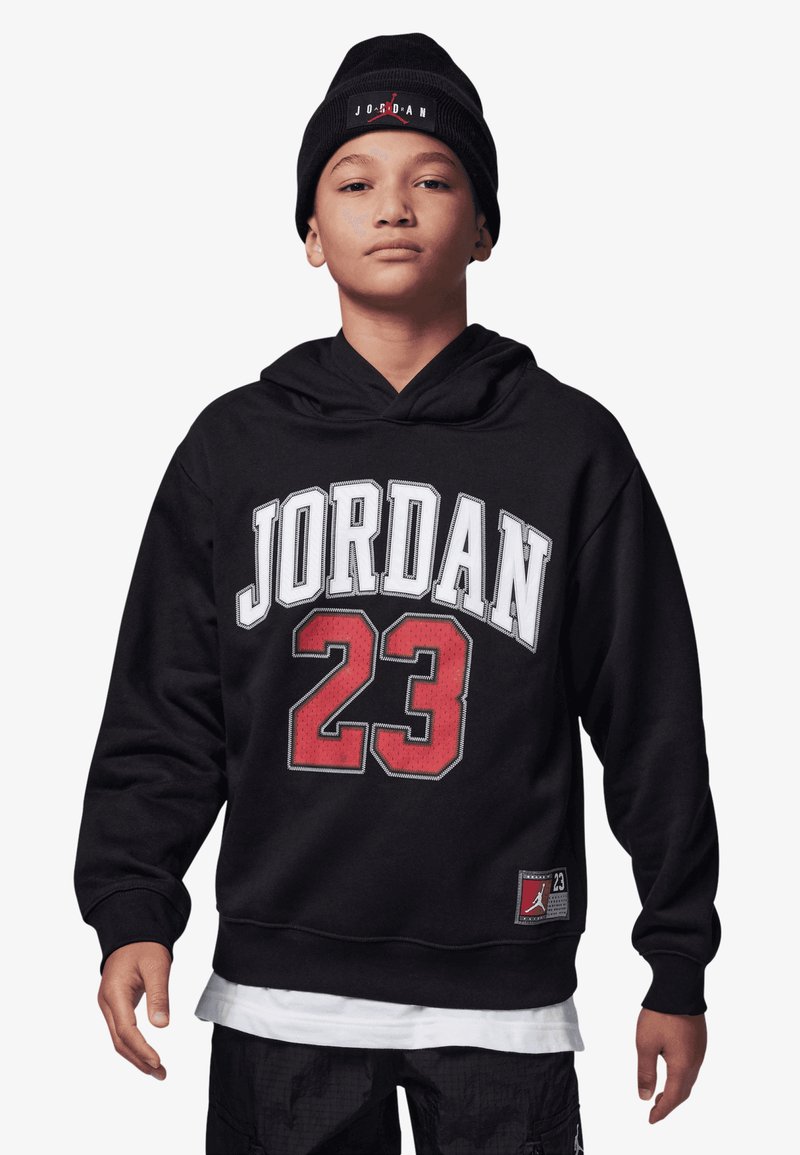 Hoodie preto com o texto branco "JORDAN" e o "23" vermelho na frente. Apresenta um bolso canguru e uma etiqueta com um logótipo na barra. Modelo usando um gorro preto.
