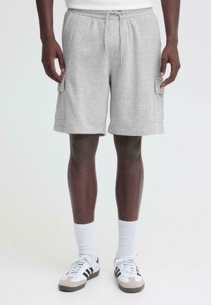 Mand iført lysegrå cargoshorts med snor i taljen, hvide ankelstrømper og hvide sneakers med sorte striber, stående foran en ensfarvet baggrund.