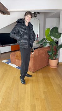 Doudoune noire avec une capuche, chemise noire à manches longues et pantalon noir. Le sujet se tient sur un sol en bois près d'un canapé et d'une plante.