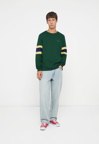 Green long-sleeve shirt with yellow and navy stripes on sleeves, paired with light blue jeans and red sneakers. Simple design, casual fit.