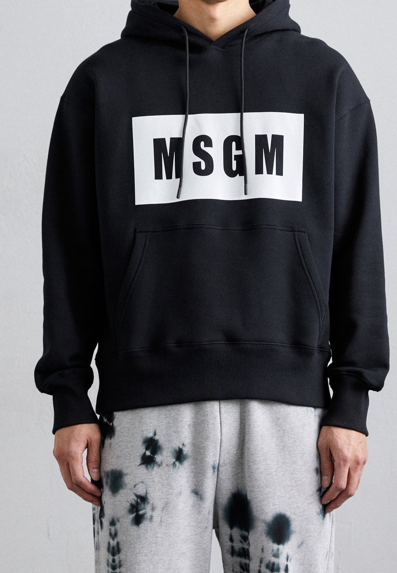 Personne portant un sweat à capuche MSGM noir avec un rectangle logo blanc et un pantalon de jogging gris à motifs tie-dye noirs, debout contre un mur uni.