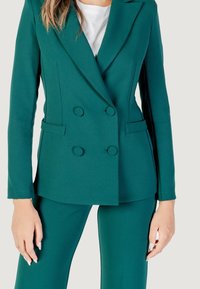 Mujer lleva un blazer ceñido color teal con botones de doble pecho, combinado con pantalones a juego, fotografiada contra un fondo neutro.