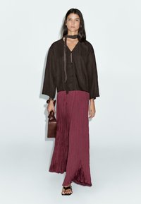 Blouse ample marron foncé avec un détail à nouer, associée à une jupe maxi plissée bordeaux. Le mannequin tient un sac à main en cuir marron.