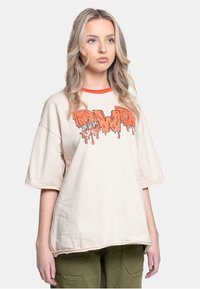 Beige oversized t-skjorte med oransje dryppende grafisk trykk; har rund hals med kontrasterende rød kant og korte ermer.
