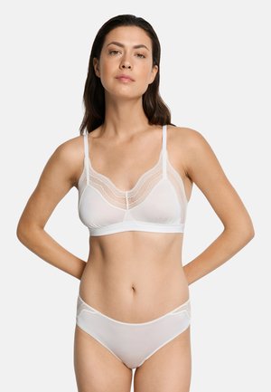 Kvinde med langt mørkt hår iført hvid bh med blonder og matchende trusser, stående med hænderne bag ryggen foran en ensfarvet baggrund.