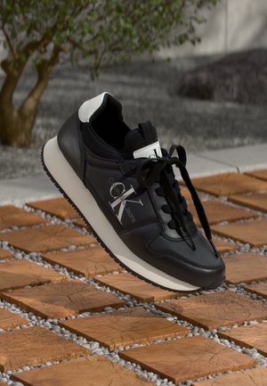 Sneaker in pelle nera con dettagli bianchi, lacci rotondi, suola in gomma testurizzata e logo laterale. Elevato sopra una superficie decorata.