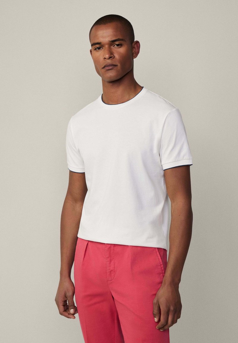 T-shirt bianca in cotone con scollo rotondo e maniche corte, con una rifinitura blu navy sul collo e sulle maniche, indossata con pantaloni rosa.