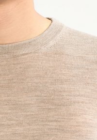 Hellbeiger Pullover mit Rundhalsausschnitt, geripptem Kragen und feiner Strickstruktur. Das Material wirkt weich und leicht glänzend.