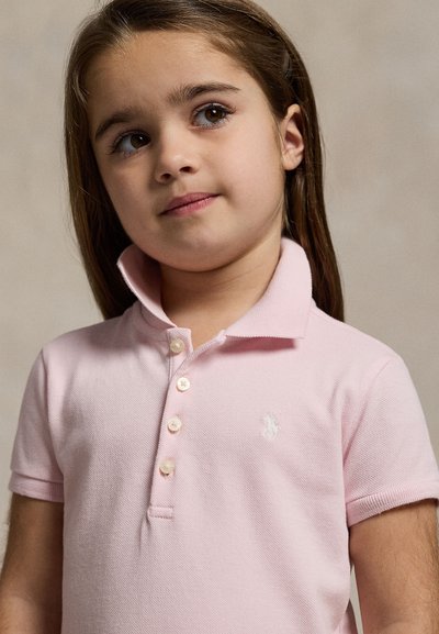 Junges Mädchen mit langen braunen Haaren, das ein hellrosa Poloshirt trägt und leicht zur Seite schaut mit neutralem Gesichtsausdruck.