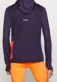 Sudadera atlética de manga larga morada con un diseño texturizado, un bolsillo frontal en naranja y detalles reflectantes, llevada sobre leggings naranjas.