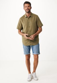 Olivgrön kortärmad skjorta med knappar, denimshorts och vita sneakers. Man som står med händerna ihop mot en neutral bakgrund.