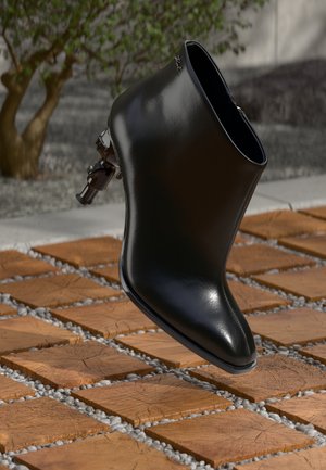 Bota de tornozelo em verniz preto com bico fino e elegante, fecho lateral com zíper e detalhes em metal, exibida sobre um fundo de azulejos de madeira.