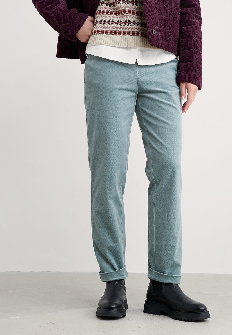 Personne portant un pantalon en velours côtelé bleu clair à jambes droites, des bottes noires massives, un pull superposé, une chemise et une veste violette, debout sur un sol gris.