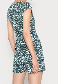 Grüner, floraler Jumpsuit mit kurzen Ärmeln, tailliertem Oberteil, gebundenem Bund und Shorts. Das Muster besteht aus kleinen weißen Blumen auf dunklem Hintergrund.