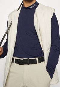 Närbild av en person som bär en marinblå långärmad skjorta med RLX-logotyp, beige väst, beige byxor, svart bälte, och håller en golfklubba över axeln.