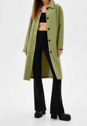 Femme portant un long manteau vert olive sur un haut noir court, un pantalon noir évasé taille haute et des chaussures à plateforme noires, sur fond blanc.