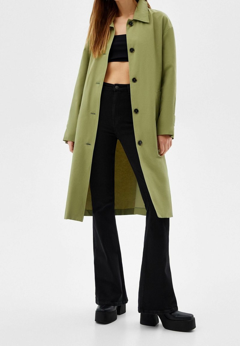 Femme portant un long manteau vert olive sur un haut noir court, un pantalon noir évasé taille haute et des chaussures à plateforme noires, sur fond blanc.