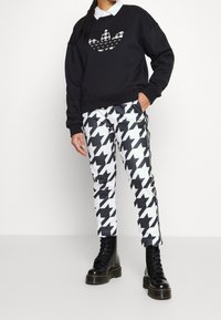 Černá mikina se vzorem houndstooth, doplněná bílými kalhotami s černým houndstooth vzorem a dvěma postranními pruhy. Černé šněrovací boty.