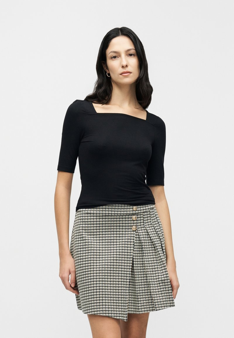 Μαύρο εφαρμοστό τοπ με τετράγωνο ντεκολτέ και κοντά μανίκια, συνδυασμένο με φούστα σε σχέδιο καρό houndstooth με πιέτες μπροστά και λεπτομέρειες με κουμπιά.