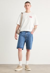 Witte t-shirt met korte mouwen met rood "Calvin Klein" logo, blauwe denim shorts, beige sneakers, witte sokken en een lichtbruine schoudertas.