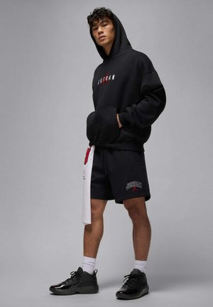 Sudadera negra con el logo rojo de "JORDAN", combinada con pantalones cortos negros que tienen un logo. Zapatillas negras y calcetines blancos completan el conjunto.