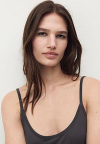 Gray tank top made of a smooth fabric with thin straps, featuring a scooped neckline and a minimalist design. Hair is dark and slightly wavy.