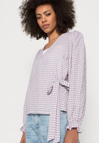 Top envolvente em padrão xadrez rosa e branco, com mangas longas bufantes e detalhe de laço na cintura, apresentando um tecido texturizado e punhos com franzido.