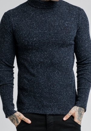 Man draagt een nauwsluitende donkergrijze geribbelde coltrui met geborduurd logo op de borst en heeft zijn handen gedeeltelijk in de zakken van een zwarte broek.