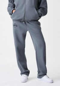 Calças de moletom cinzas feitas de material suave, com um cós elástico e um logótipo impresso dos Los Angeles Lakers no lado esquerdo.