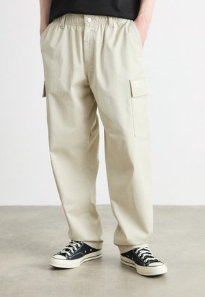 Pantalones cargo beige claro con cintura elástica y bolsillos laterales, combinados con zapatillas negras con cordones blancos y suelas de goma.