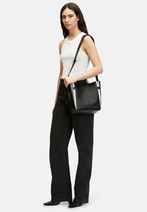Black leather tote bag with white accents, structured shape, and adjustable strap, displayed alongside a person wearing a sleeveless top and black pants.