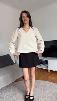 Frau steht in einem Wohnzimmer, trägt eine beige langärmelige Bluse, einen schwarzen Rock und schwarze Schuhe, mit einem Fernseher und Sofa im Hintergrund.