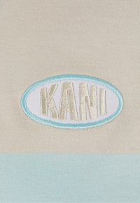 Le tissu en coton présente un patch oval brodé avec "KANI" en lettres dorées claires sur un fond beige avec un accent bleu clair.
