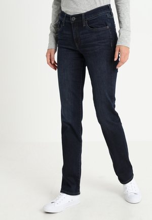 Kvinde iført mørkeblå jeans med lige ben, lysegrå langærmet top og hvide sneakers, stående mod en ensfarvet baggrund.