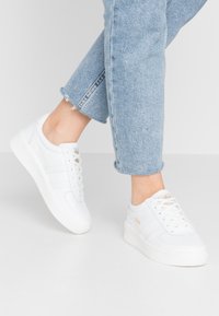 Sneakers bianchi in pelle con superficie testurizzata, dotate di suola in gomma e un discreto accentuazione del logo sul lato. Abbinati a jeans in denim blu.