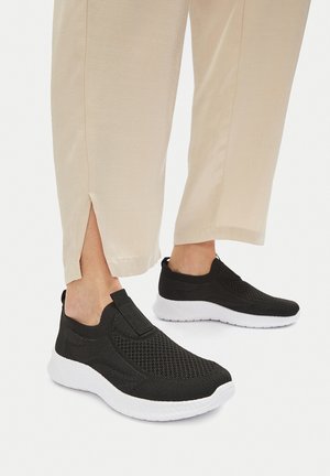 Schwarze Mesh-Slips mit gepolstertem Kragen und weißer Sohle, kombiniert mit beigefarbenen, knöchellangen Hosen mit seitlichen Schlitzen.