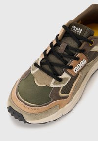 Zapatilla marrón, verde y beige con materiales de malla y ante, forma redondeada, cordones negros y un prominente logo de Colmar en el lateral.