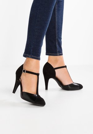 Ben i mørkeblå jeans iført sorte T-rem hæle med spænder mod en hvid baggrund.