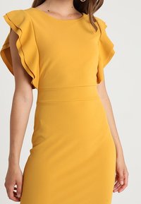 Gul klänning med volangärmar, figurnära midja och en slät textur. Enkel halsringning, knälång design. Inga synliga mönster.