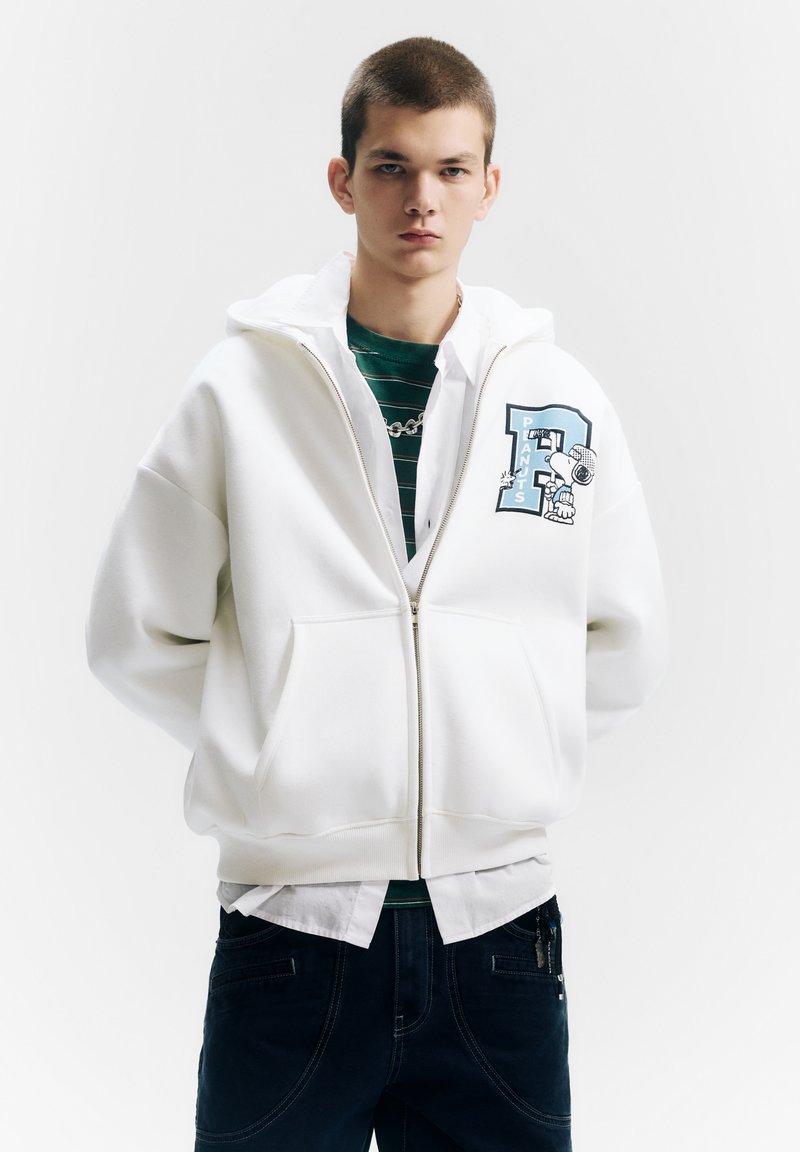 Witte hoodie met ritssluiting en grote zakken aan de voorkant, met een blauw grafisch ontwerp op de borst, gecombineerd met een groen gestreept shirt en donkere broek.