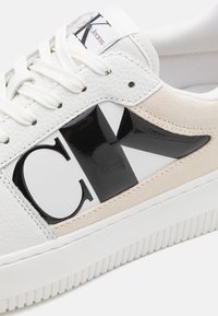 Vit lädersneakers med svart och vit logodesign, beige mockadetaljer och strukturerad gummisula med ränder. Snörning är vit.