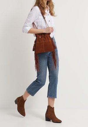 Sac seau en daim marron avec détails en franges, fermeture à cordon et sangle réglable. Porté avec un jean court bleu clair et des bottines marron.